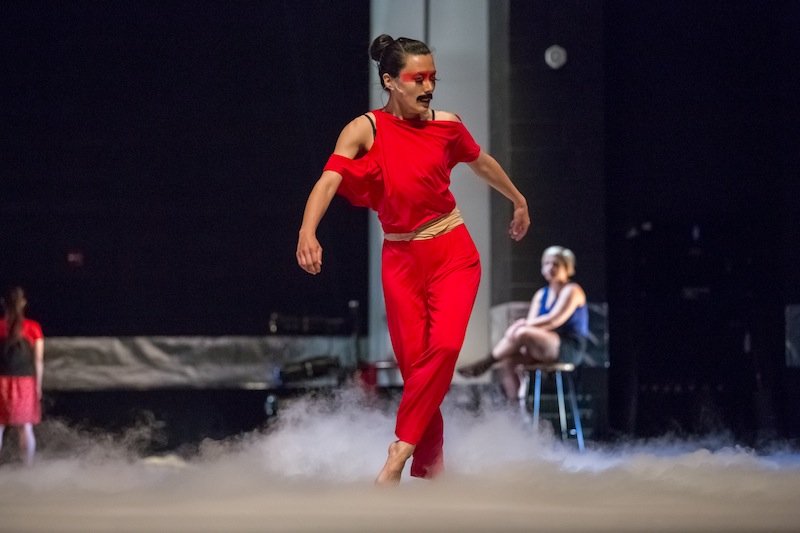A female dancer wearing a bright red two piece pant costume. Smoke billows at her feet. She has red warrior paint around her eyes